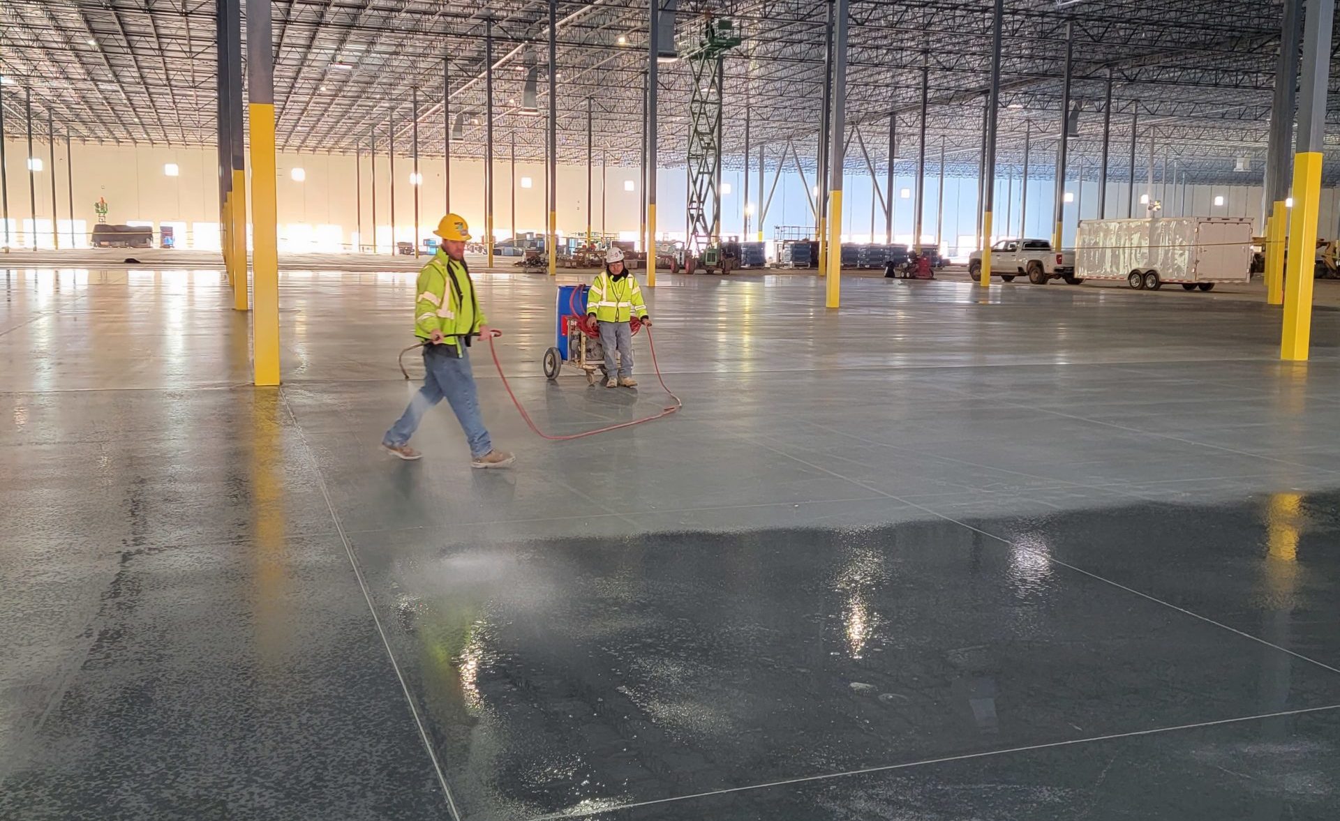 Two men in yellow safety jackets perform floor work in a large industrial shell.